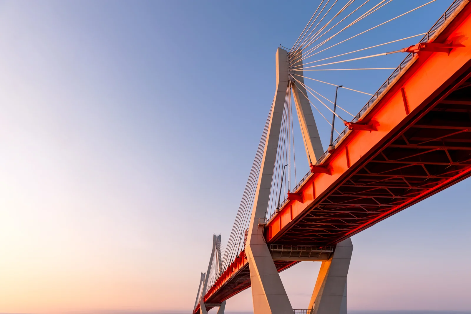 A red cable-stayed bridge at sunset stretching across a calm sea — a metaphor for the connections Deltara structures between international principals and regional markets