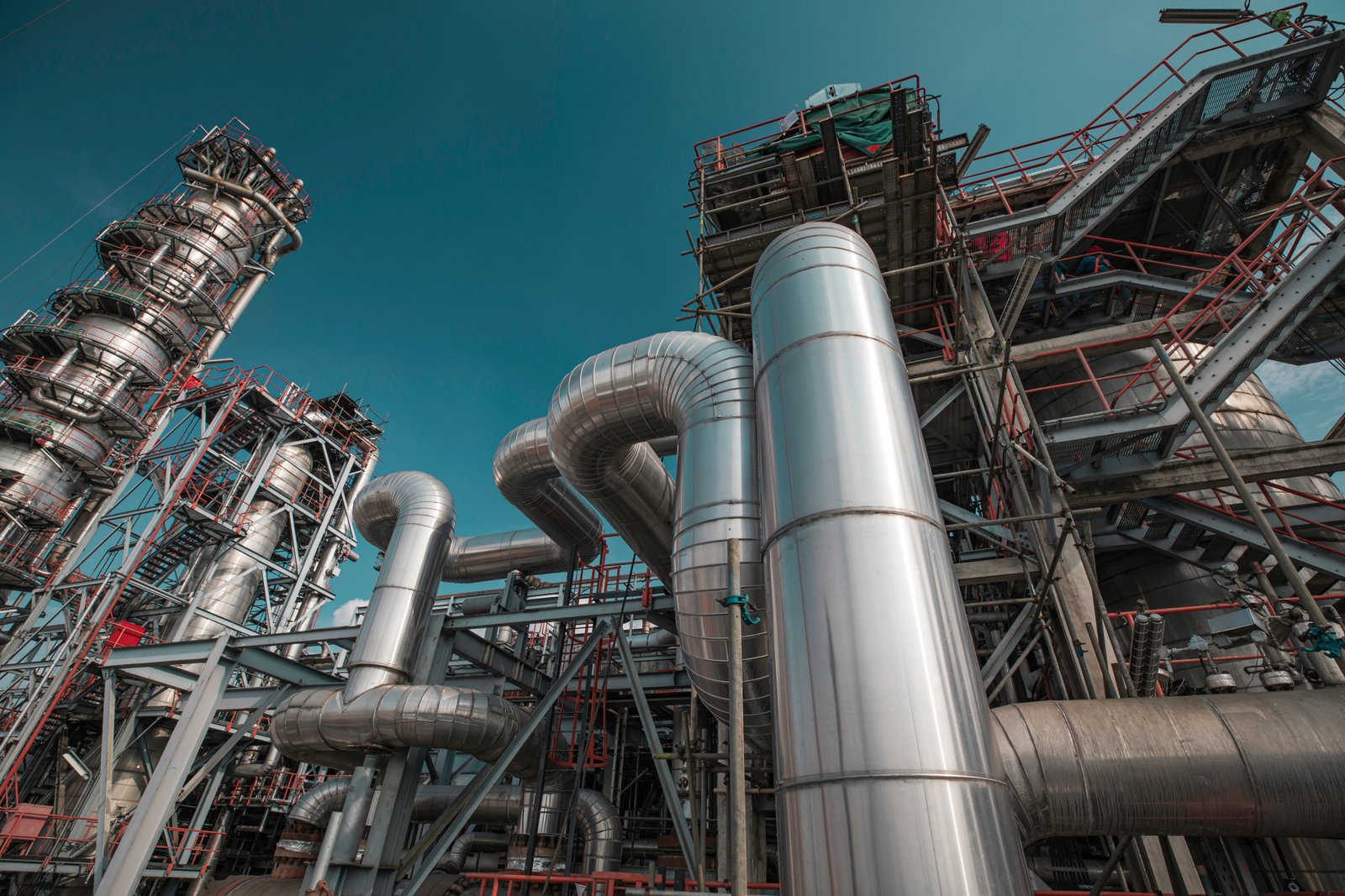 Bright daylight industrial refinery equipment with large silver pipes and red structural framework